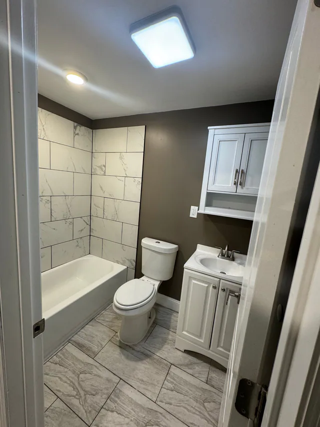 Finished bathroom with marble-look tub surround, white tub, toilet, vanity, and wall cabinet