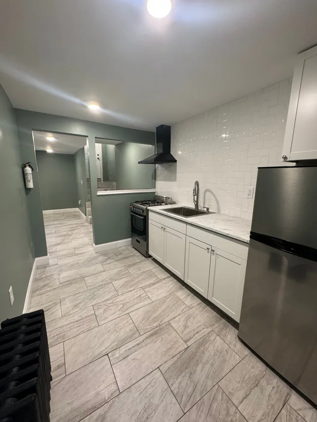 Finished galley kitchen with white cabinets, marble counter, black range hood, range, refrigerator, and wood-look tile floor