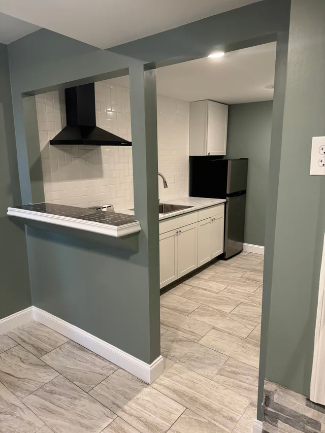 Finished kitchen seen through a serving pass-through, with white shaker cabinets, black range hood, white subway tile, stainless refrigerator, and wood-look tile floor